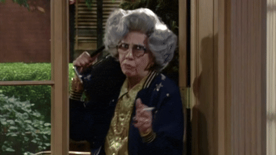 Zia Yetta, l'anziana ed eccentrica zia della sitcom La Tata, con i suoi iconici occhiali spessi e i capelli cotonati, mentre fuma la sua immancabile sigaretta con un'espressione placida e del tutto indifferente al caos che la circonda